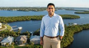 Smiling man in a blue shirt standing by a scenic waterfront in Jupiter, Florida, with residential properties and lush greenery in the background, representing local property management insights.